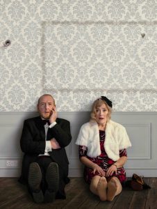 Two actors sat on the floor against the wall looking to camera