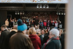 Photo of the stage at the lights switch on from a year gone by. Man is on the stage with a mic speaking to the crowd