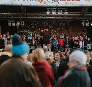 Photo of the stage at the lights switch on from a year gone by. Man is on the stage with a mic speaking to the crowd