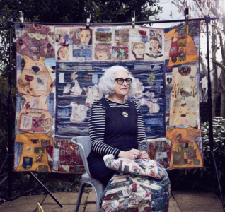 An artist sat with her works on a chair. She's sat outside looking into the distance with her works on a board behind her.
