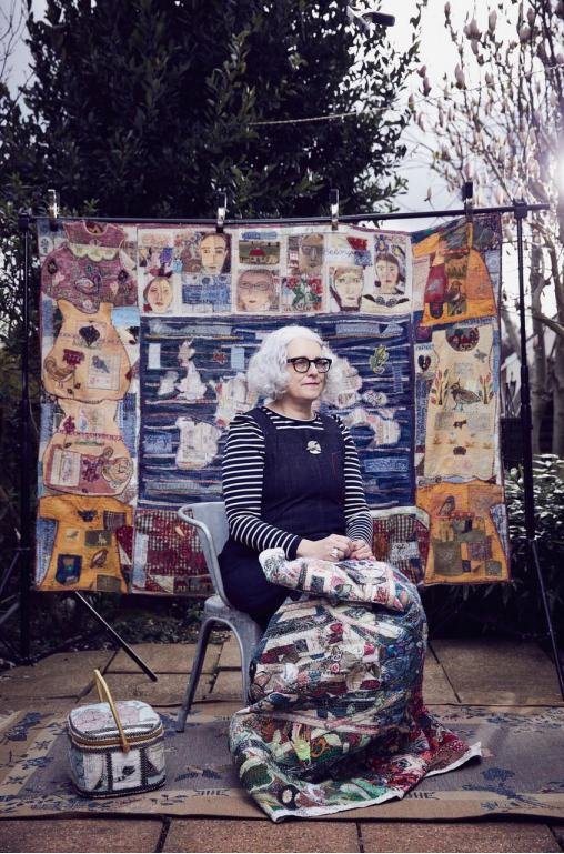 An artist sat with her works on a chair. She's sat outside looking into the distance with her works on a board behind her.