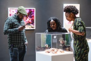 Three people looking at an art exhibition in a small perspex box in an art gallery