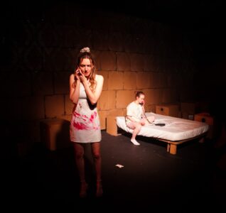 Actor on the stage in blood splattered clothes with someone in the background of the stage sat on the bed.