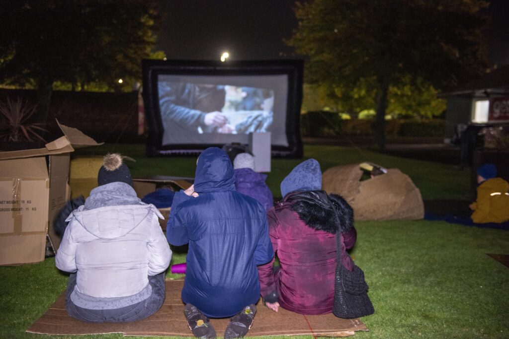 Three people sat on cardboard outside in a park watching a big screen