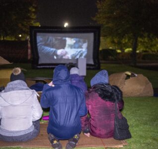 Three people sat on cardboard outside in a park watching a big screen