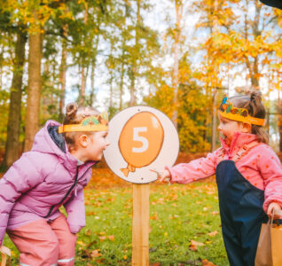 Two young girls stood by a sign in the forest with an easter egg pained on it with the number 5.