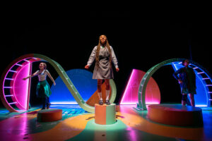 Three young students on stage, one of them in the centre is in the spotlight and on a pedestal.