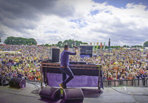 The image shows Pat Sharp from the back standing on stage in front of a large crowded audience.