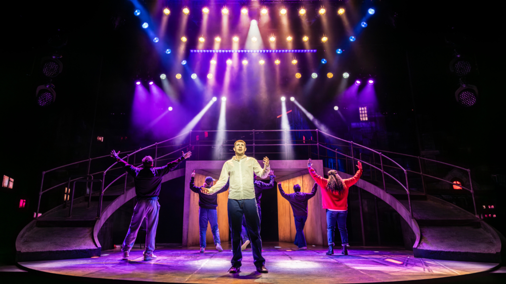 A stage set with sweeping ramps down both sides and strong spotlights, in shades of blue, pink and purple. 6 actors fill the stage. All but one face the back, their arms raised. The man at the front holds his arms out, as if asking a question.