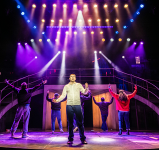 A stage set with sweeping ramps down both sides and strong spotlights, in shades of blue, pink and purple. 6 actors fill the stage. All but one face the back, their arms raised. The man at the front holds his arms out, as if asking a question.